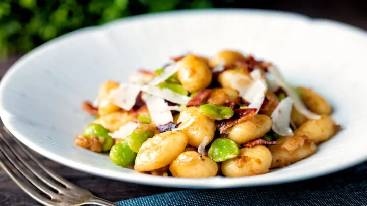 Pan fried gnocchi with bacon, broad beans and parmesan shavings served in a white bowl.