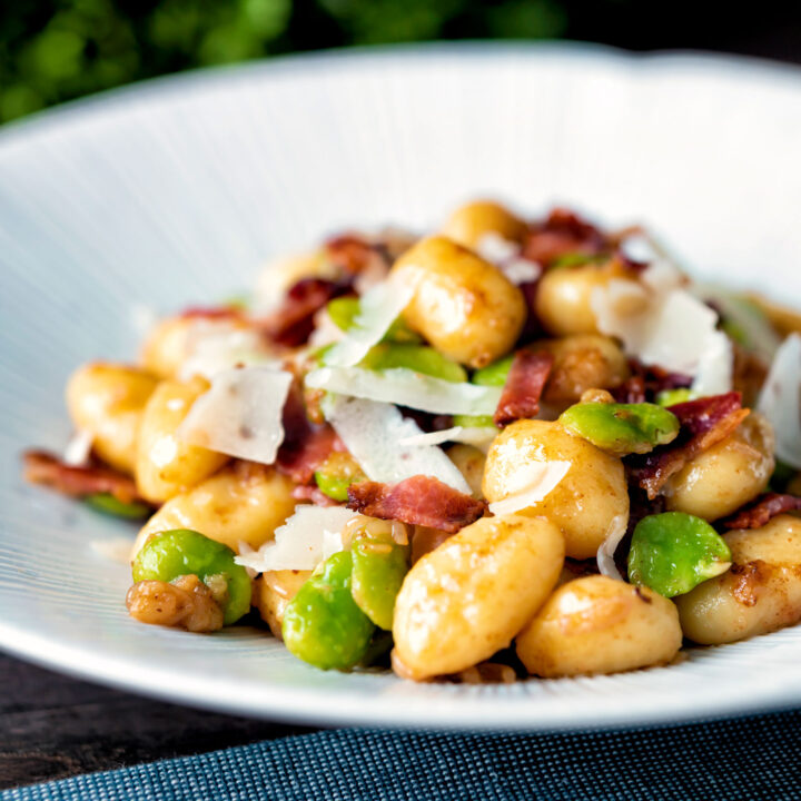 Pan fried gnocchi with bacon, broad beans and parmesan shavings served in a white bowl.