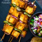 Overhead paneer tikka skewers served with kachumber salad and naan bread featuring a title overlay.