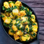 Overhead aloo methi or fenugreek potatoes served in a black dish featuring a title overlay.