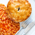 Overhead corned beef and potato pie wrapped in shortcrust pastry served with baked beans featuring a title overlay.