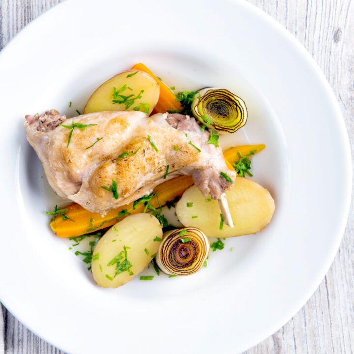 Braised rabbit legs served with leeks, carrots and potatoes in a white bowl.