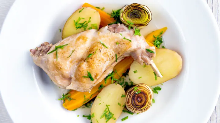Braised rabbit legs served with leeks, carrots and potatoes in a white bowl.