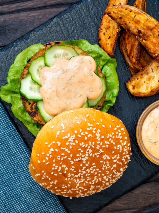 Overhead spicy cod fish burger with sriracha mayonnaise served with potato wedges.