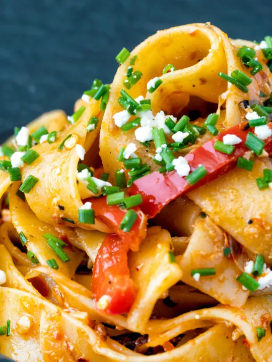 Close up harissa pasta with with feta cheese and roasted peppers in a black bowl.