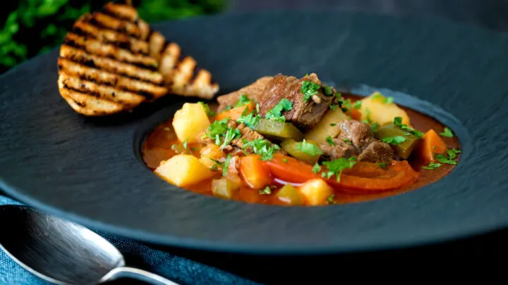 Hungarian lamb goulash soup served with griddled bread in a black bowl.