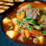 Close up Hungarian lamb goulash garnished with parsley served in a black bowl featuring a title overlay.