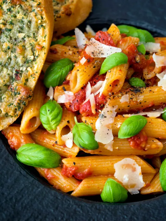 Close up Italian penne arrabbiata served with basil, parmesan, black pepper and cheesy garlic.