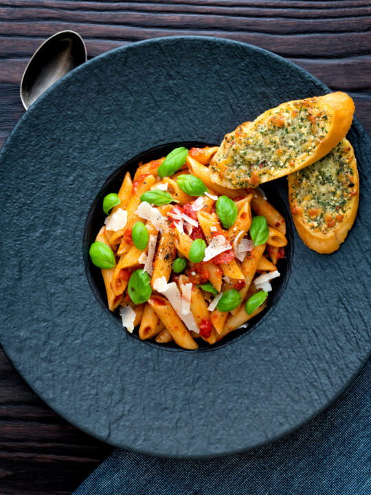 Overhead Italian penne arrabbiata served with basil, parmesan, black pepper and cheesy garlic bread.