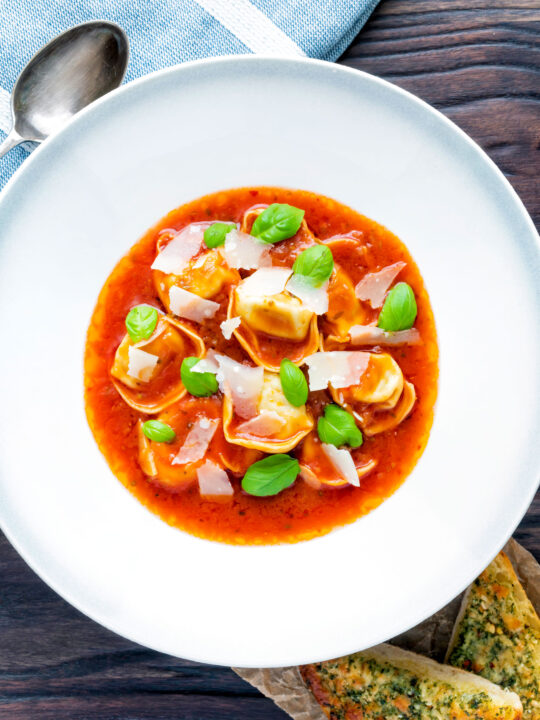 Overhead store bought tortellini soup with tomato, fresh basil and parmesan shavings.