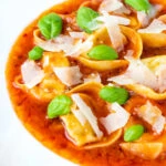 Close up store bought tortellini soup with tomato, fresh basil and parmesan shavings.