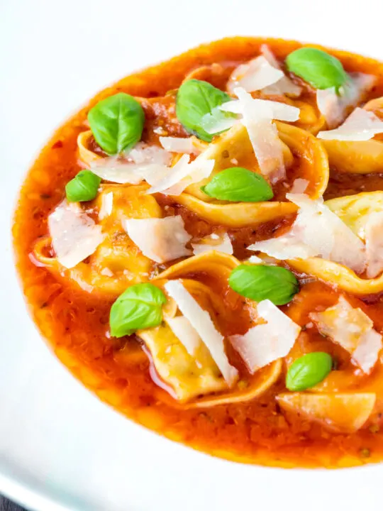 Close up store bought tortellini soup with tomato, fresh basil and parmesan shavings.