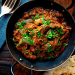 Overhead lamb bhuna curry served in an iron karahi with naan bread featuring a title overlay.