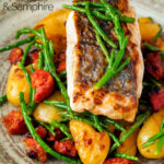 Close up pan fried hake with chorizo, new potatoes and samphire featuring a title overlay.