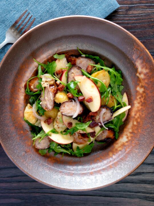 Overhead sausage salad with apple, new potatoes, bacon and rocket or arugula.