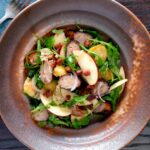 Overhead sausage salad with apple, new potatoes, bacon and rocket or arugula featuring a title overlay.