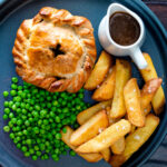 Overhead hand raised steak and ale pie with chips and peas with a title overlay.