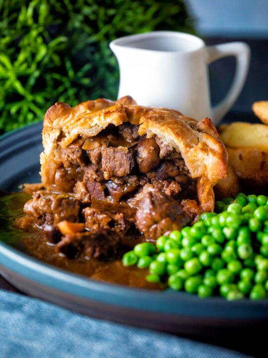 Hand raised steak and ale pie cut open to show filling served with chips and peas.
