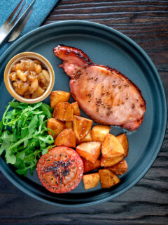 Overhead cider glazed bacon chops with fried potatoes, grilled tomatoes and rocket.