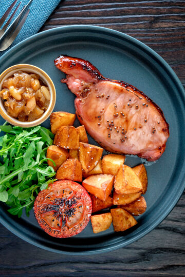 Overhead cider glazed bacon chops with fried potatoes, grilled tomatoes and rocket.