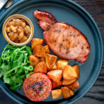 Overhead cider glazed bacon chops with fried potatoes, grilled tomatoes and rocket featuring a title overlay.