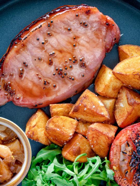 Overhead close up cider glazed bacon chops with fried potatoes, grilled tomatoes and rocket.