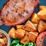 Overhead close up cider glazed bacon chops with fried potatoes, grilled tomatoes and rocket featuring a title overlay.