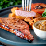 Cider glazed cut open bacon chops with fried potatoes, grilled tomato, rocket and apple chutney featuring a title overlay.