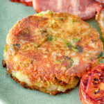Close up fried bubble and squeak patty served with bacon, grilled tomato and fried egg.