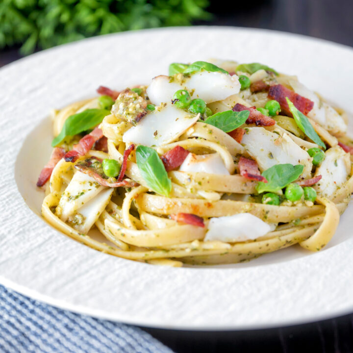 Cod fish pasta with pesto, peas and bacon served on a white plate.