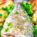Close up baked or roasted sea bass with new potatoes, peas, broad beans and bacon featuring a title overlay.