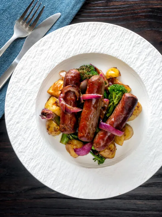 Overhead sausage and potato tray bake with onions and broccoli served on a plate.