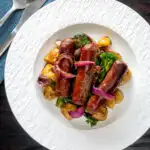 Overhead sausage and potato tray bake with onions and broccoli served on a plate featuring a title overlay.