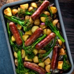 Overhead sausage and potato tray bake with onions and tenderstem broccoli.