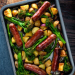Overhead sausage and potato tray bake with onions and tenderstem broccoli featuring a title overlay.