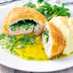 Golden chicken Kyiv cut open showing herby garlic butter filling served with mashed potato.
