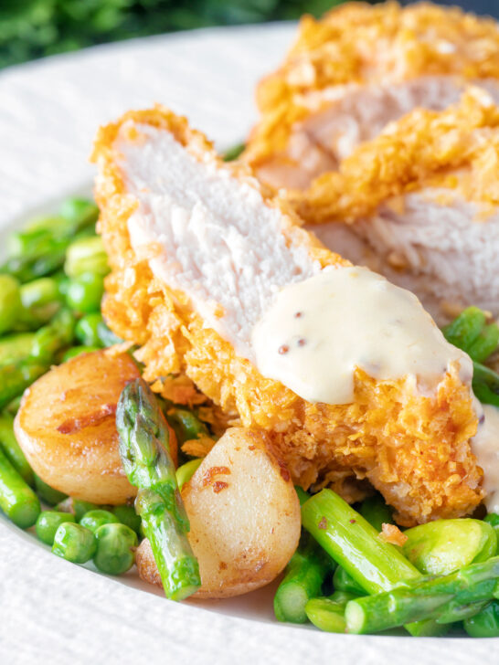 Close up oven baked cornflake coated chicken breast served with mustard sauce and veggies.