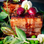Close up twice cooked pressed crispy pork belly with port cherries and peas featuring a title overlay.