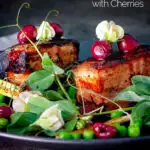 Twice cooked pressed crispy pork belly with port cherries and peas featuring a title overlay.