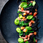 Overhead bacon pasta with squid ink spaghetti, spinach and tomatoes featuring a title overlay.