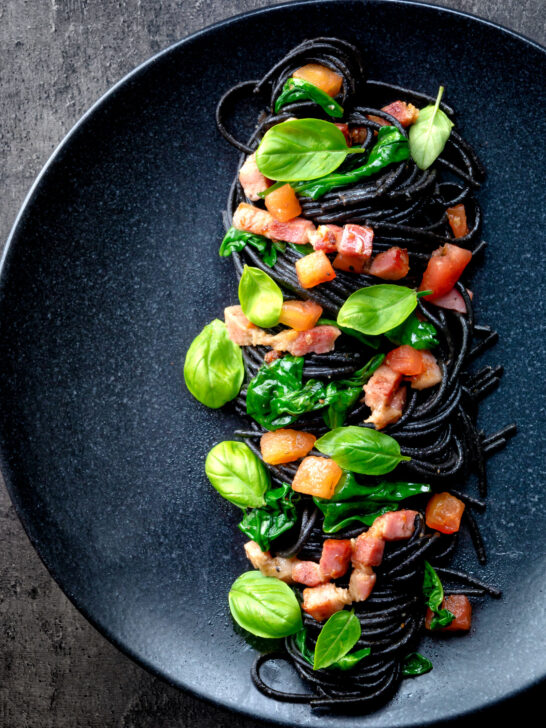 Overhead bacon pasta with squid ink spaghetti, spinach and tomatoes featuring a title overlay.