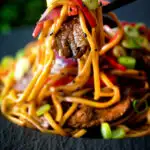 Beef noodle stir fry with sweet chilli sauce and red peppers being eaten with chopsticks featuring a title overlay.