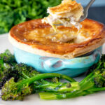Puff pastry topped individual chicken and leek pie showing creamy filling served with broccoli.