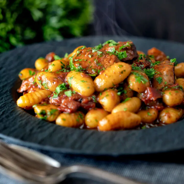 Steaming hot portion of sausage gnocchi in a tomato sauce on a black plate.