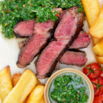 Overhead close-up rare cooked rump steak with chimichurri sauce and chips.
