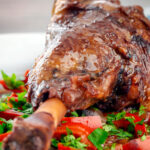 Close up slow cooker braised lamb shanks in a red wine, tomato, garlic and pepper sauce featuring a title overlay.