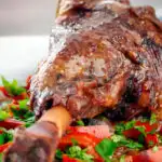 Close up slow cooker braised lamb shanks in a red wine, tomato, garlic and pepper sauce featuring a title overlay.