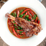 Overhead slow cooker braised lamb shanks in a red wine, tomato, garlic and pepper sauce.
