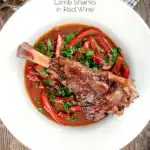Overhead slow cooker braised lamb shanks in a red wine, tomato, garlic and pepper sauce featuring a title overlay.