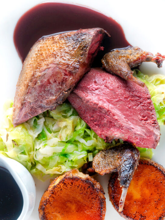 Overhead close-up roast pigeon breasts, confit legs, red wine sauce, buttered cabbage and fondant potatoes.